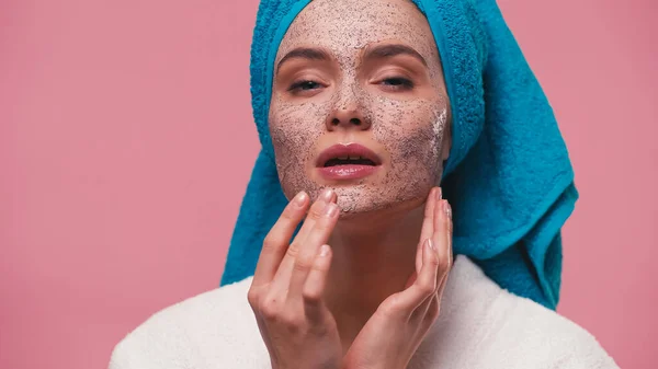 Mujer con toalla azul en la cabeza aplicando exfoliante cosmético en la cara aislado en rosa - foto de stock