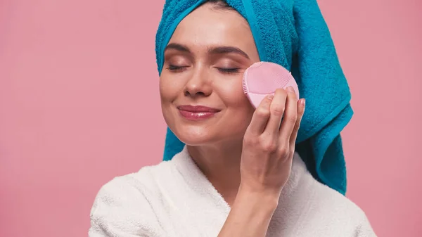 Femme heureuse avec les yeux fermés en utilisant une brosse nettoyante faciale en silicone isolée sur rose — Photo de stock