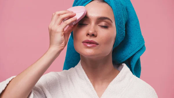 Mujer con los ojos cerrados limpiando la cara con cepillo de silicona eléctrica aislado en rosa - foto de stock