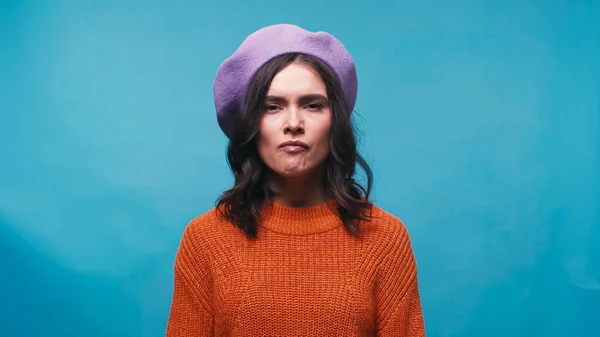 Angry woman in beret and jumper looking at camera isolated on blue — Stock Photo