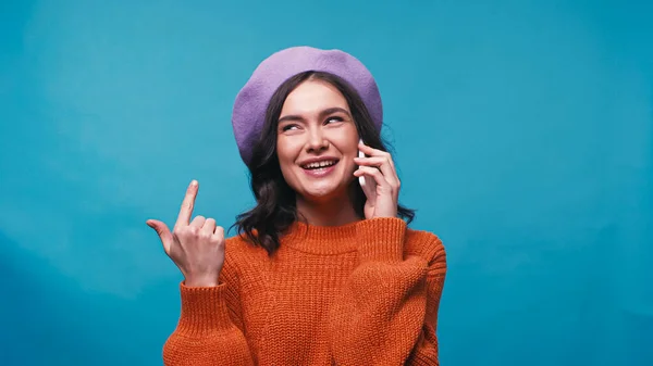 Mujer alegre en boina lila apuntando a sí misma mientras habla en el teléfono celular aislado en azul - foto de stock