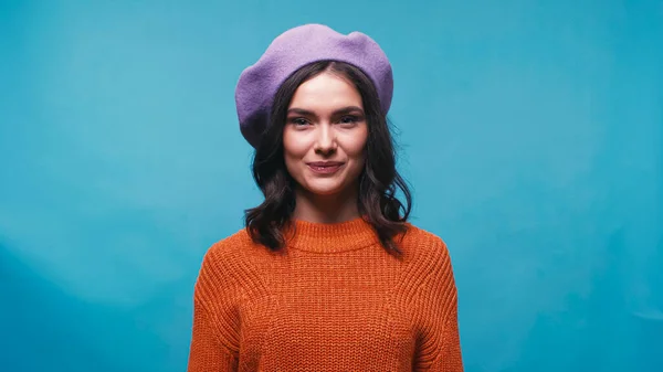 Brunette woman in knitted sweater and beret smiling at camera isolated on blue — Stock Photo