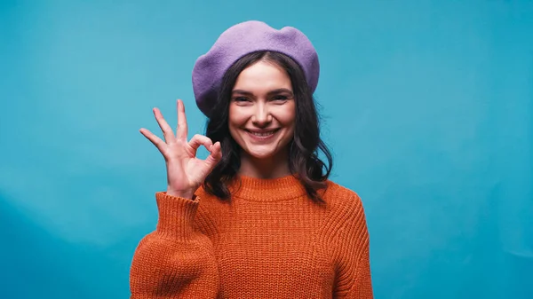 Positive junge Frau in fliederfarbener Baskenmütze zeigt okay Geste isoliert auf blau — Stockfoto