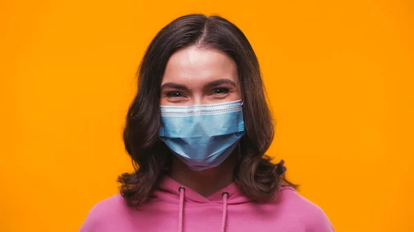 Positive brünette Frau in medizinischer Maske schaut in die Kamera isoliert auf gelb — Stockfoto