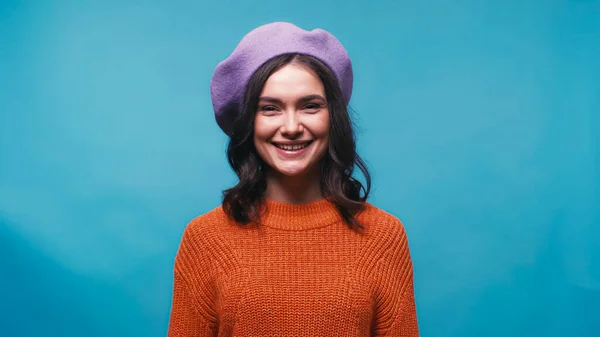 Mujer alegre en suéter de punto y boina lila sonriendo a la cámara aislada en azul - foto de stock