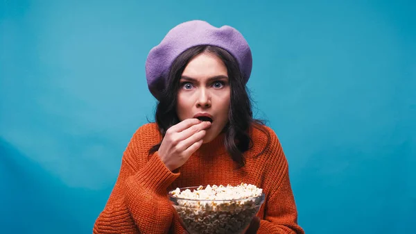 Mujer preocupada en boina púrpura viendo película y comiendo palomitas aisladas en azul - foto de stock