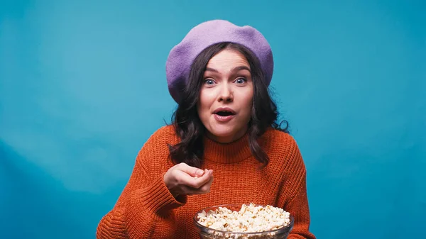 Mulher maravilhada em jumper e boina assistindo filme emocionante e comer pipoca isolada no azul — Fotografia de Stock