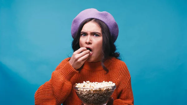 Besorgte Frau in fliederfarbener Baskenmütze isst Popcorn, während sie Film isoliert auf blau anschaut — Stockfoto