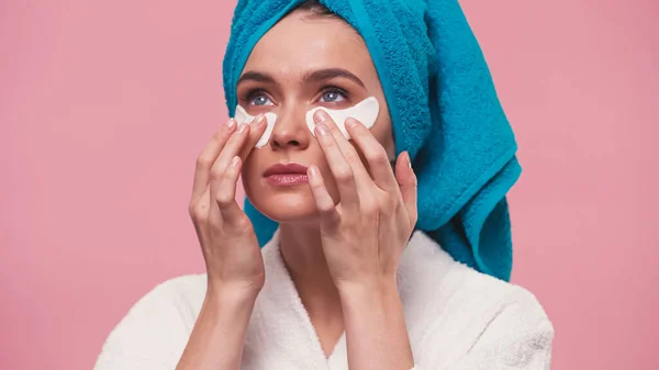 Giovane donna con asciugamano blu sulla testa applicando macchie oculari isolate su rosa — Foto stock