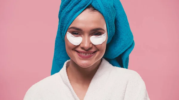 Pleased woman with eye patches and towel on head smiling at camera isolated on pink — Stock Photo