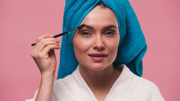 Young woman with natural makeup brushing eyebrows isolated on pink — Stock Photo