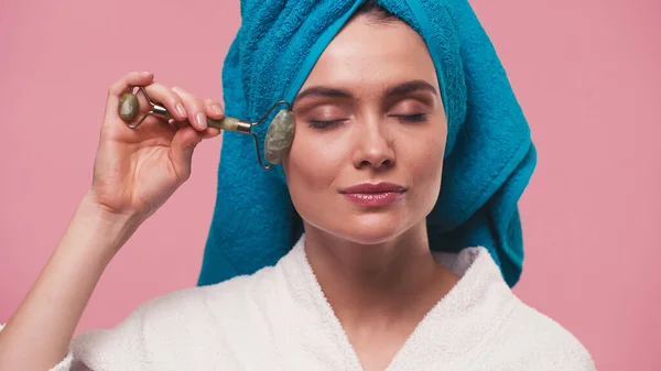 Young woman with towel on head massaging face with jade roller isolated on pink — Stock Photo