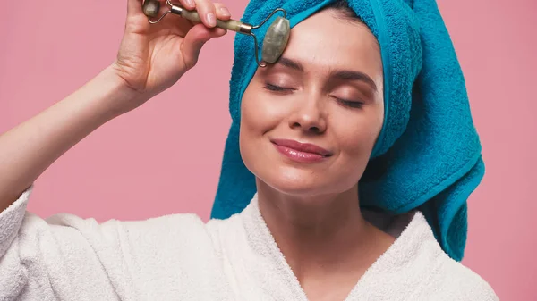 Donna soddisfatta con occhi chiusi massaggio viso con rullo di giada isolato su rosa — Foto stock