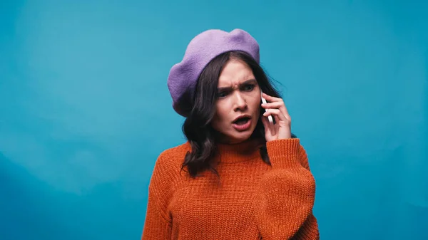 Angry woman in sweater and beret talking on mobile phone isolated on blue — Stock Photo