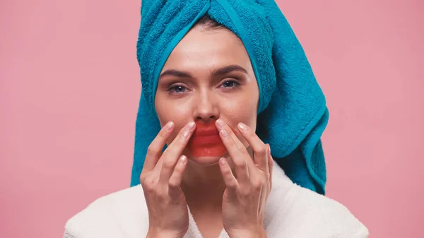 Young woman with towel on head looking at camera and applying lip mask isolated on pink — Stock Photo