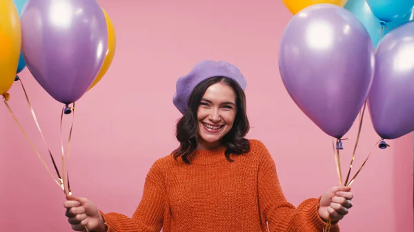 Mujer alegre en boina y jersey sosteniendo globos multicolores aislados en rosa - foto de stock