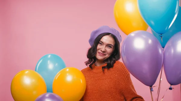 Mulher feliz em boina e suéter sorrindo perto de balões festivos isolados em rosa — Fotografia de Stock