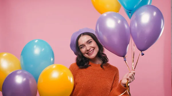 Fröhliche Frau in warmem Pullover und Baskenmütze lächelt in die Kamera neben bunten Luftballons auf rosa — Stockfoto