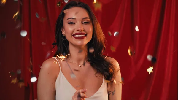 Mujer alegre con tatuaje celebración brillante chispeante cerca de confeti en rojo - foto de stock