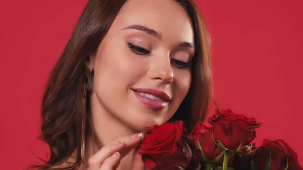 Feliz joven tocando rosas aisladas en rojo - foto de stock