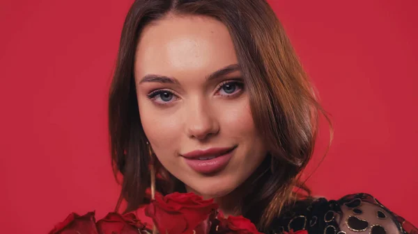 Bonita joven sonriendo cerca de rosas aisladas en rojo - foto de stock