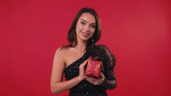 Freudige junge Frau lächelt, während sie eine Geschenkschachtel auf Rot hält — Stockfoto
