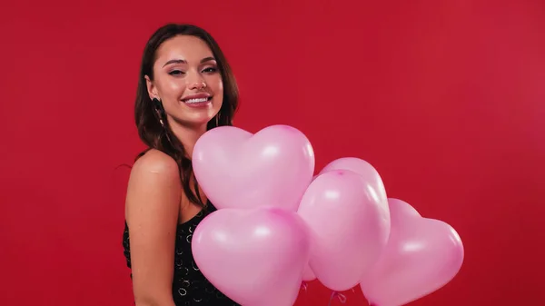 Mulher feliz em vestido preto segurando balões rosa isolado no vermelho — Fotografia de Stock