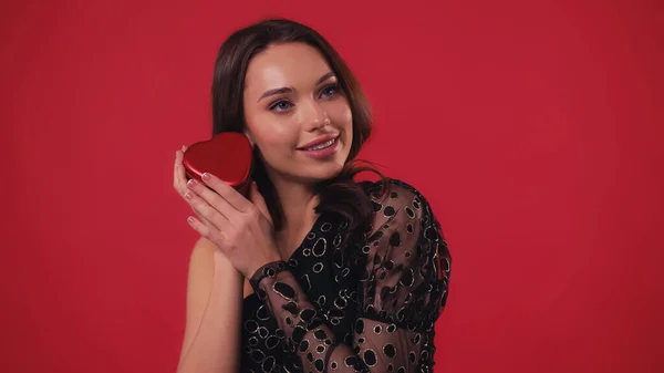 Alegre mujer sosteniendo en forma de corazón caja de regalo aislado en rojo - foto de stock