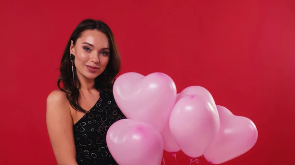 Cheerful woman in black dress holding pink balloons isolated on red — Stock Photo