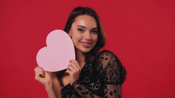Young woman holding paper heart and smiling isolated on red — Stock Photo