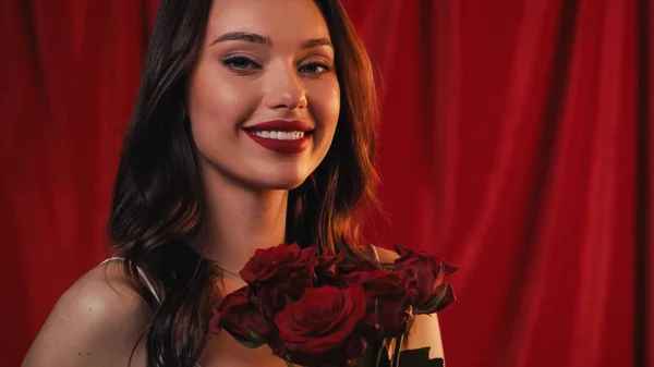 Щаслива молода жінка з яскравими губами посміхається біля троянд на червоному — стокове фото