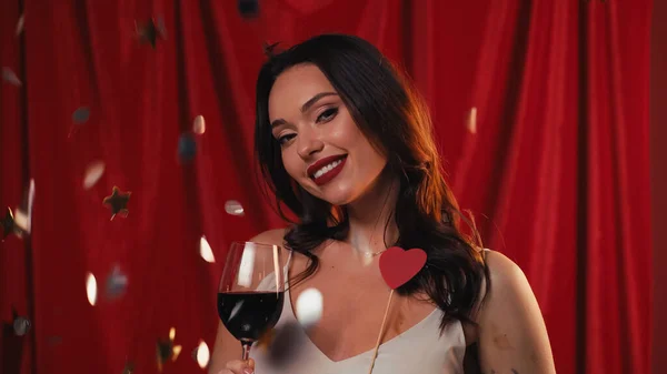 Happy woman holding glass of wine near falling confetti on red — Stock Photo