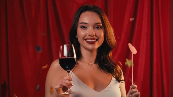 Cheerful woman holding glass of wine and heart on stick near falling confetti on red — Stock Photo