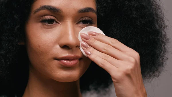 Nahaufnahme einer afrikanisch-amerikanischen Frau, die ihr Gesicht mit einem Wattepad isoliert auf grau wischt — Stockfoto