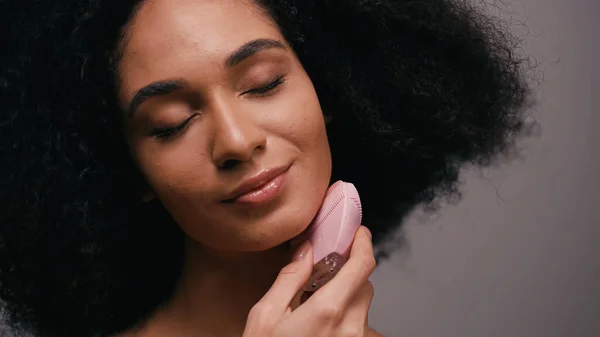 Mujer afroamericana complacida usando limpiador de silicona en la cara aislada en gris - foto de stock