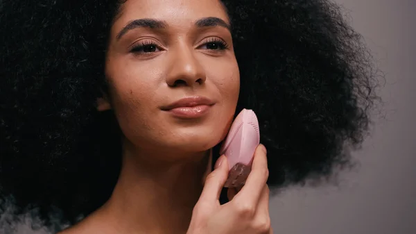 Кучерява афроамериканська жінка використовує силіконовий засіб для чищення на обличчі ізольовано на сірому — стокове фото