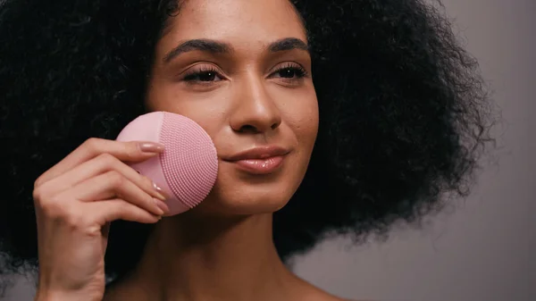 Brunette african american woman using silicone cleanser on face isolated on grey — Stock Photo