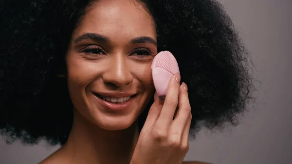 Happy african american woman using silicone cleansing device on face isolated on grey — Stock Photo