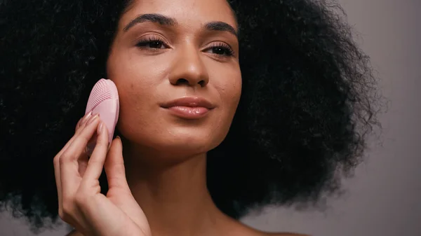 Кучерява афроамериканська жінка використовує силіконовий пристрій для очищення обличчя ізольовано на сірому — стокове фото