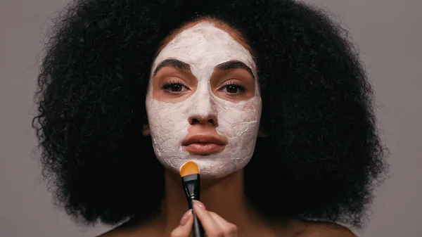 Mulher afro-americana olhando para a câmera e aplicando máscara de barro isolado em cinza — Fotografia de Stock