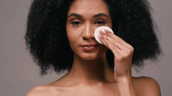 Mujer afroamericana joven limpiando la cara con almohadilla de algodón aislado en gris - foto de stock