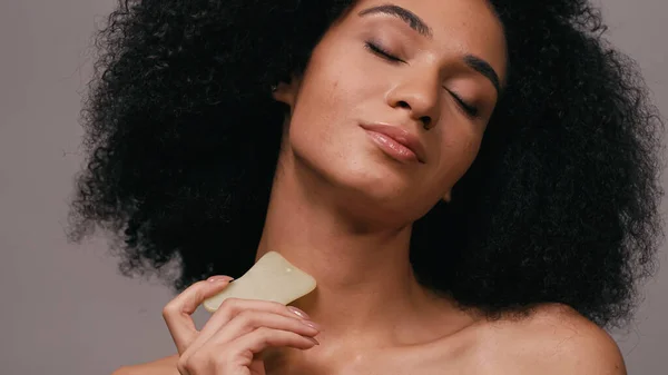 Curly african american woman with closed eyes using gua sha stone isolated on grey — Stock Photo