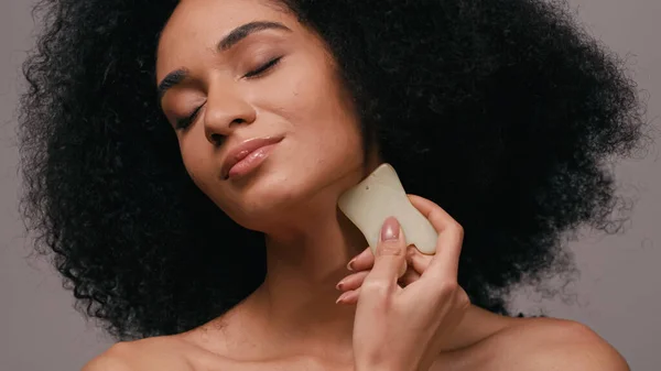 Brunette african american woman with closed eyes using gua sha stone isolated on grey — Stock Photo