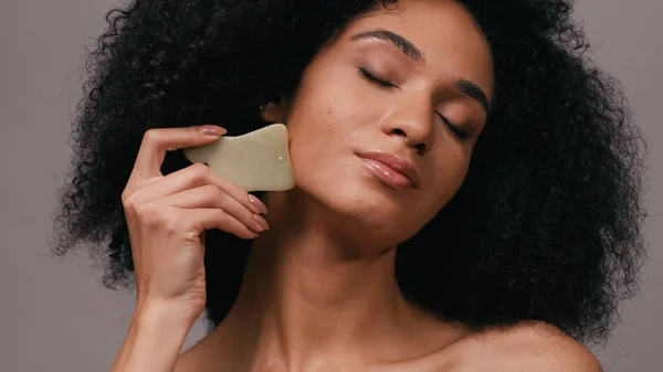 Jovem afro-americana com olhos fechados usando gua sha pedra isolada em cinza — Fotografia de Stock