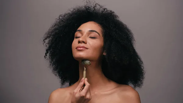 Curly african american woman with bare shoulders using jade roller isolated on grey — Stock Photo