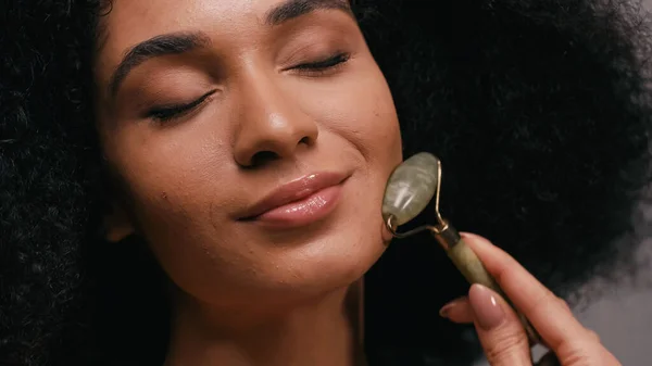 Close up of pleased african american woman with closed eyes using jade roller — Stock Photo