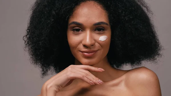 Sonriente mujer afroamericana con crema cosmética en la cara aislada en gris - foto de stock