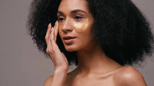 Mujer americana africana rizada aplicando parches para los ojos de colágeno aislados en gris - foto de stock