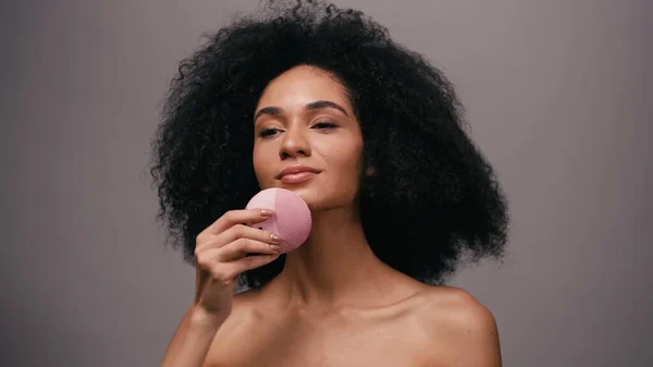 Young african american woman with bare shoulders using silicone cleansing device on face isolated on grey — Stock Photo