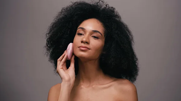 Femme afro-américaine aux épaules nues utilisant un dispositif de nettoyage en silicone sur le visage isolé sur gris — Photo de stock